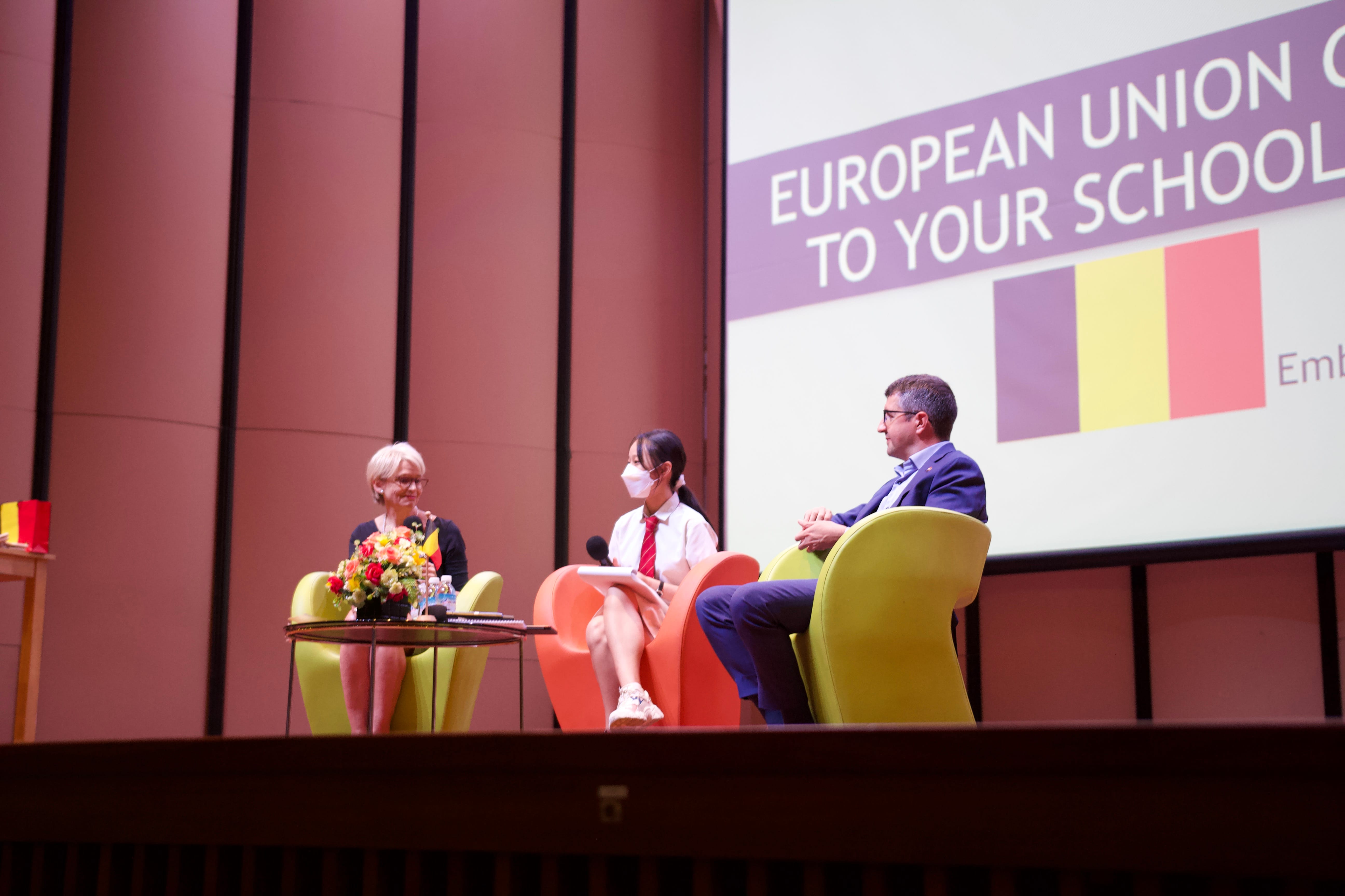 Three people sit on colorful chairs on a stage with a projected slide reading "European Union Comes to Your School."